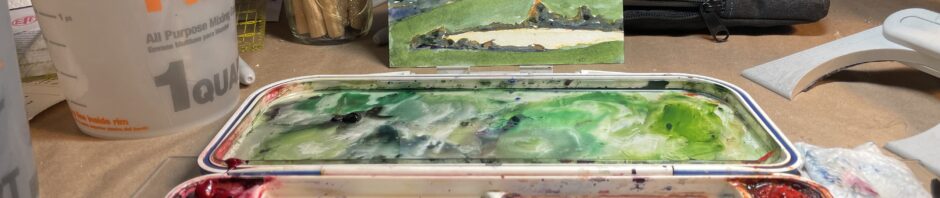 foreground has my watercolor palette with small slots of colors around outer edge and three big mixing areas. Background has a container of water, my jar of lino-cutting tools and, center, a postcard done by printing a block in yellow and using watercolor to fill in areas.