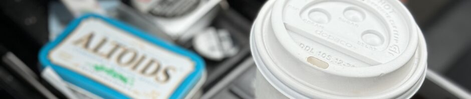 White take out coffee cup with brown sleeve and white lid sits in car cupholder with assorted car flotsam: altoids tin and measuring tape.