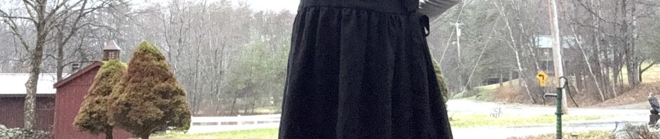 Going off to Easter brunch in my new dress. Woman standing in driveway wearing black dress with a white long sleeve shirt underneath, red tights, a black bowler with a big red flower. Her right outstretched arm is held at shoulder height.
