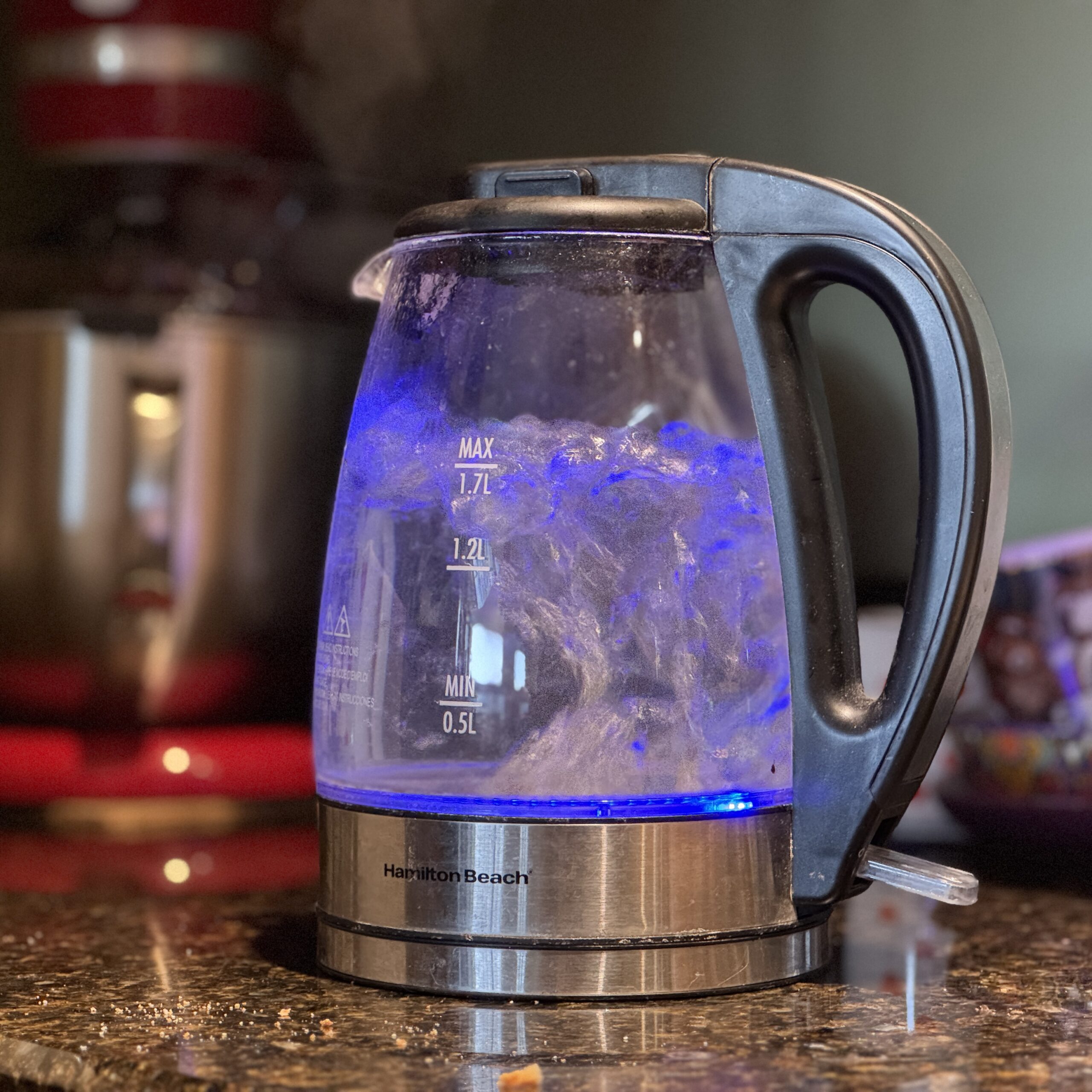 Photo: a glass electric water kettle is sitting on a counter with a mixer in the background. The kettle has a black handle and lid. The water inside is beginning to boil and is lit from the kettle base with a blue light.