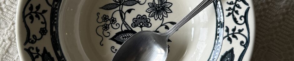 A small bowl with a blue onion pattern around the rim and center has a stainless steel spoon with a beaded edge resting in it. They both sit on a handwoven cream-color cloth.