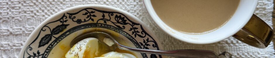 photo: on a natural color hand-loomed placemat, a white bowl with blue design along edge holds several chopped up eggs and some butter. Nearby is a big white mug of coffee, regular. In between sits a folded pair of glasses.