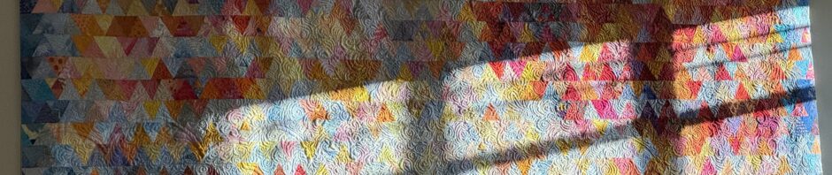 Photo of my quilt "A Place to Stand" hanging on the wall with light from the door's window across it. The quilt is made of small equilateral triangles in yellows, oranges and blues in an abstract design.