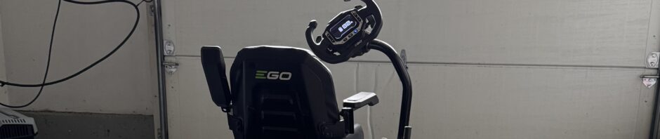 Photo: Electric ride-on mower sits in a garage to charge. The garage door is closed and light comes in through two windows that are curved at the top and have multiple panes. The mower is facing toward the door. The control screen indicates 82% charge and the battery indicators in the back are glowing green.