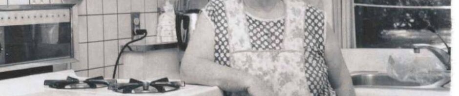 Black and white photo: an older woman with white hair stands in the center of the photo, window and sink to the right and big white gas stove and oven to the left. She leans her elbow on the edge of the stove. She's wearing an apron over a light sleeveless dress.